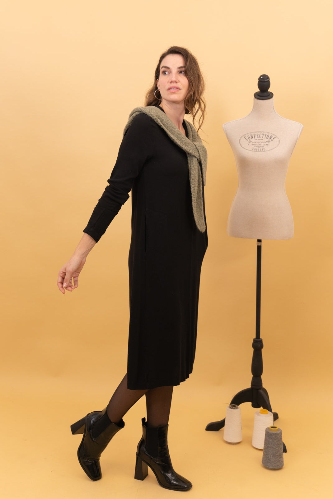 Vista lateral del vestido midi Spirit negro sobre modelo, con jersey sobre los hombros y botines de tacón